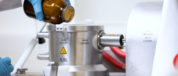 In a laboratory setting, a person wearing blue gloves carefully pours liquid from a brown bottle into a metal container, utilizing Co-Precipitation Technology to enhance flowability and improve bulk density.