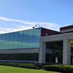 A modern brick and glass building stands proudly with a grassy lawn and trees under a clear blue sky, reminiscent of the purity sought in APIs and chemical intermediates.