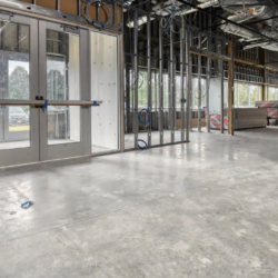 The interior of the unfinished commercial building reveals a state-of-the-art design with exposed framework, concrete floors, and sleek glass entry doors. Construction materials are visible in the background, hinting at its future as a cutting-edge facility.