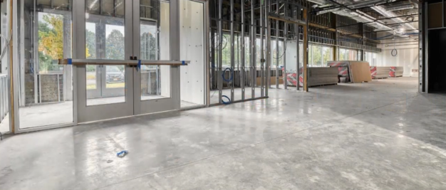 The interior of the unfinished commercial building reveals a state-of-the-art design with exposed framework, concrete floors, and sleek glass entry doors. Construction materials are visible in the background, hinting at its future as a cutting-edge facility.