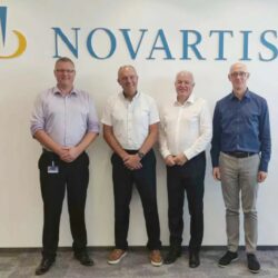 Four men stand in front of a wall adorned with the Novartis logo, poised to discuss plans to expand operations in Europe.