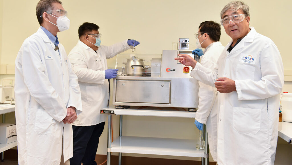 Four scientists in white lab coats and masks stand in a laboratory setting, with one pointing to equipment on a table.