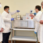 Four scientists in white lab coats and masks stand in a laboratory setting, with one pointing to equipment on a table.