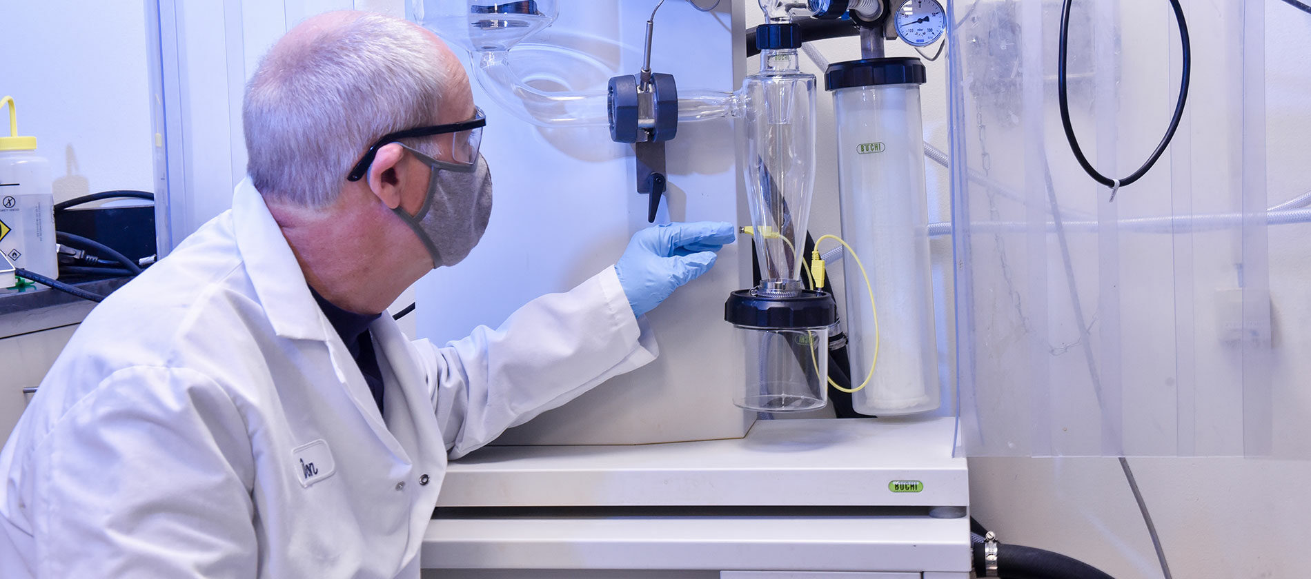 A scientist in a lab coat and mask works with laboratory equipment, adjusting controls and observing gauges and containers.