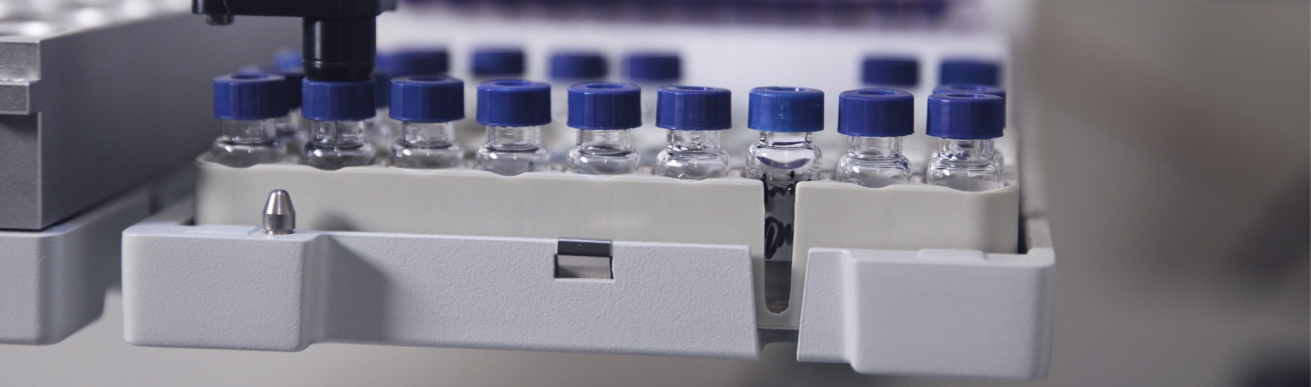 A row of small glass vials with blue caps secured in a white holder, positioned under a black mechanical arm, likely part of laboratory equipment.