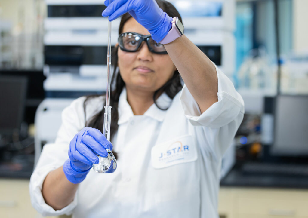 A person in a lab coat and gloves carefully pours liquid from a pipette into a flask in a laboratory setting.