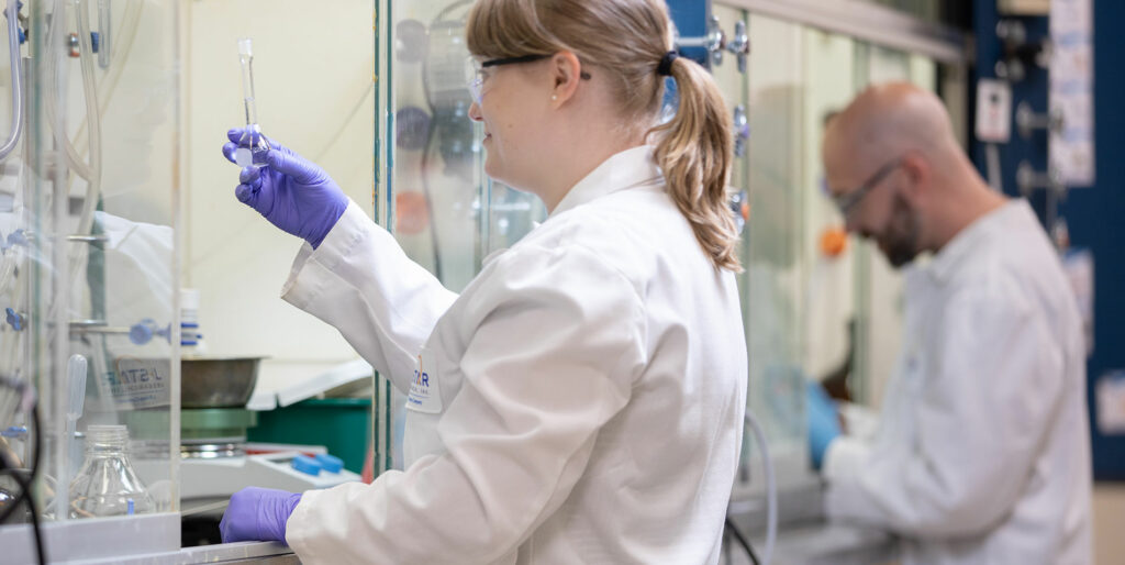 Two scientists in white lab coats and gloves working in a laboratory, with one examining a test tube and the other focused on an experiment.