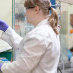 Two scientists in white lab coats and gloves working in a laboratory, with one examining a test tube and the other focused on an experiment.