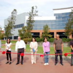 Seven people standing behind a red ribbon in front of a modern building, possibly for a ribbon-cutting ceremony.