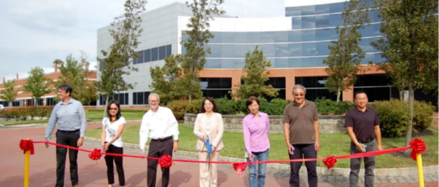 Seven people standing behind a red ribbon in front of a modern building, possibly for a ribbon-cutting ceremony.