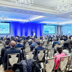 Conference attendees seated in a ballroom, facing a speaker on stage. Two screens display a presentation. The room is well-lit with chandeliers.