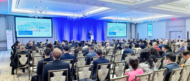 Conference attendees seated in a ballroom, facing a speaker on stage. Two screens display a presentation. The room is well-lit with chandeliers.