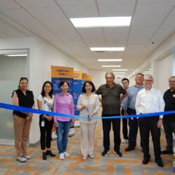 A group of people stand together in a hallway, holding a blue ribbon. A ribbon-cutting event appears to be taking place. There are banners and a sign in the background.