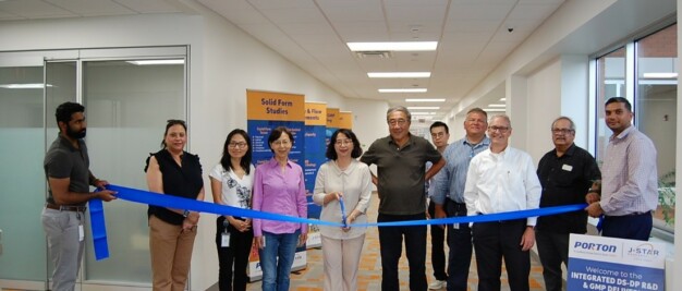 A group of people stand together in a hallway, holding a blue ribbon. A ribbon-cutting event appears to be taking place. There are banners and a sign in the background.