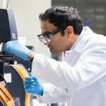 A scientist wearing protective eyewear and a lab coat operates laboratory equipment, adjusting a component with blue gloves.