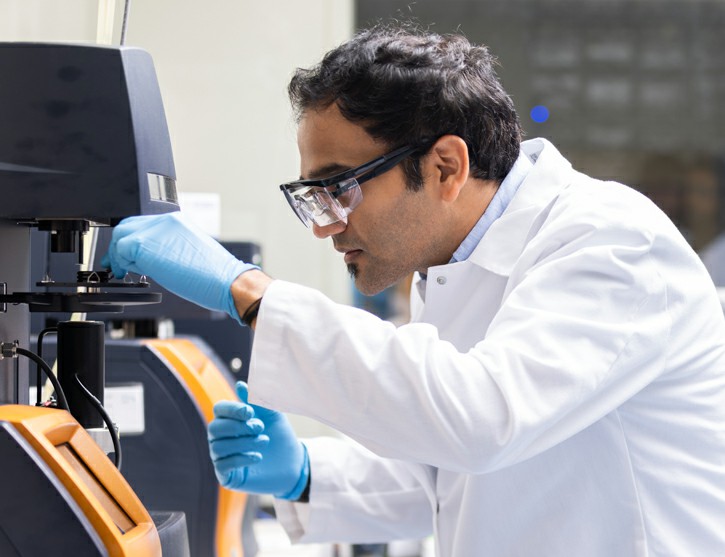 A scientist wearing protective eyewear and a lab coat operates laboratory equipment, adjusting a component with blue gloves.