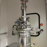 A metallic laboratory reactor with gauges and valves is positioned in a controlled environment. It is surrounded by various connecting tubes and pipes.