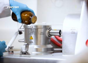 In a laboratory setting, a person wearing blue gloves carefully pours liquid from a brown bottle into a metal container, utilizing Co-Precipitation Technology to enhance flowability and improve bulk density.