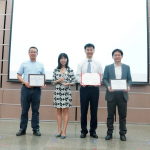 Four people proudly stand in front of a screen, holding their scholarship certificates on a stage adorned with an industrial crystallization-themed tiled wall, symbolizing innovation and achievement.