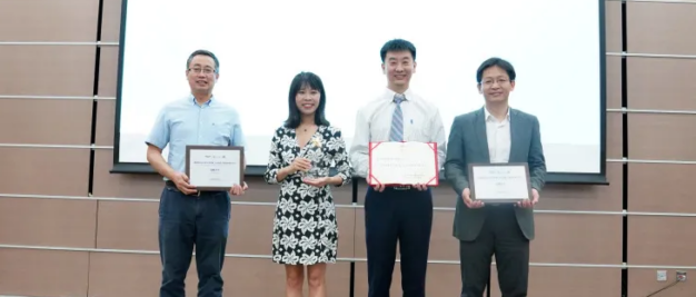 Four people proudly stand in front of a screen, holding their scholarship certificates on a stage adorned with an industrial crystallization-themed tiled wall, symbolizing innovation and achievement.