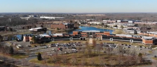 Aerial view of the CfPC's expansive office complex with multiple interconnected buildings, surrounded by parking lots, green spaces, and a pond on a clear day.