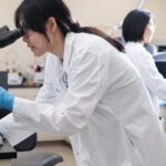 Two scientists in lab coats work in a laboratory; one examines a sample under a microscope, while the other uses a computer in the background.