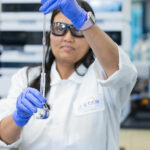 A person in a lab coat and gloves carefully pours liquid from a pipette into a flask in a laboratory setting.