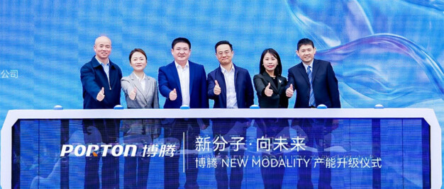 A group of six people in suits stands behind a display table with "PORTON" and Chinese text, giving thumbs up at a company event celebrating the modality capacity upgrade.