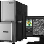 A desktop scanning electron microscope with a connected monitor displaying a highly magnified black and white image of a sample's surface texture.