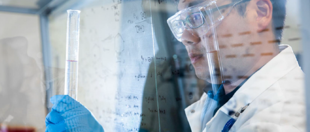 A scientist in safety glasses and gloves examines a test tube in a laboratory, where chemical equations on a glass board guide the journey of innovative drugs from preclinical to commercialization.