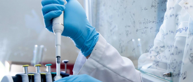 A scientist in blue gloves uses a pipette in a lab focused on pharmaceutical development, with scientific formulas and molecular modeling diagrams adorning the whiteboard in the background.